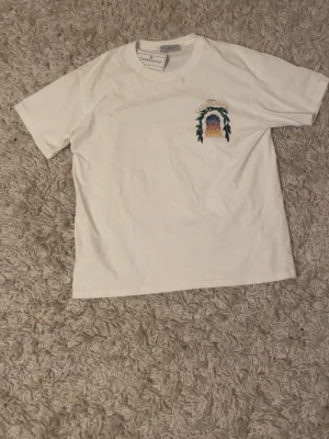 Vit Casablanca t-shirt med tryck - Vit t-shirt från Casablanca med färgglatt tryck av ett fönster och blad på bröstet och ryggen. Stort motiv på ryggen med texten 'Avenida Casablanca'. Klassisk passform och rund halsringning.
