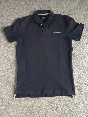 Mörkblå piké - Snygg mörkblå pikétröja med klassisk krage och korta ärmar. Tröjan har knappar vid halsen och en liten broderad logga på bröstet. Perfekt till jeans eller shorts för en avslappnad stili
