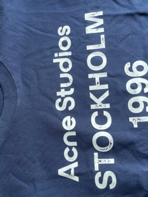 Blå Acne Studios t-shirt med tryck - Snygg blå t-shirt från Acne Studios med stort vitt tryck 'STOCKHOLM 1996' på framsidan. Klassisk rund halsringning och rak passform. Perfekt för dig som gillar stilrena och trendiga plagg med statement-tryck.