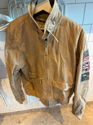 Ralph Lauren - vintage Bombarjacka  - Säljer en beige bomberjacka med hög krage och dragkedja framtill. Jackan har två stora fickor med lock och ribbade muddar. På ena ärmen finns patchar med amerikanska och brittiska flaggan samt ett runt märke. Materialet är slitstarkt bomullstyg.
