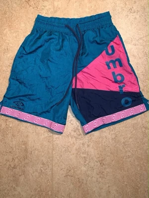 Retroblå shorts från Umbro XS - Säljer ett par retroblå shorts från Umbro med färgglada detaljer i rosa och mörkblått. Shortsen har elastisk midja med snörning och grafiskt mönster längs kanten. Perfekta för sommarens alla aktiviteter!