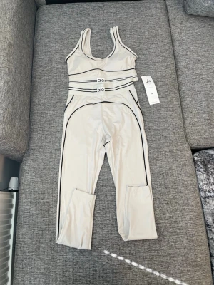 Beige träningsset från Alo Yoga - Snyggt beige träningsset från Alo Yoga med svarta kontrastsömmar. Setet består av en sport-bh och matchande tights i stretchigt funktionsmaterial. Perfekt för yoga, gym eller annan träning. Elastisk midja med Alo-logga och modern design.