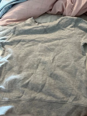 Ljusgrå sweatshirt utan huva - En basic ljusgrå sweatshirt i mjukt bomullsmaterial. Tröjan har rund halsringning, långa ärmar och ribbade muddar vid ärmslut och nederkant. Perfekt för en avslappnad och enkel stil.