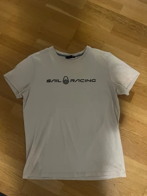 Sail Racing Tröja - Snygg beige t-shirt från Sail Racing med svart logga tryckt över bröstet. Klassisk rund halsringning och korta ärmar. Perfekt till jeans eller shorts för en clean och sportig look.