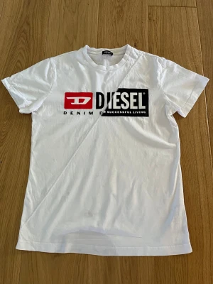 Vit Diesel t-shirt med logga - Snygg vit t-shirt från Diesel med stor logga framtill i svart och rött. Klassisk rund hals och korta ärmar. Tillverkad i mjuk bomull som känns skön mot huden. Perfekt för dig som gillar streetwear och vill ha en enkel men stilren look. Är i storlek S och är i perfekt skick