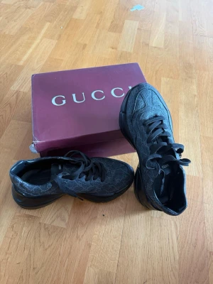 Svarta Gucci sneakers med mönster - Snygga svarta sneakers från Gucci med diskret GG-mönster över hela skon. Skorna har en chunky sula och är tillverkade i läder med textilinslag. Klassisk låg modell med svarta skosnören och detaljer. Perfekta för dig som vill ha något exklusivt och trendigt.