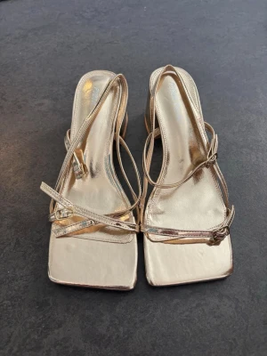 Guldiga sandaler med klack och remmar - Snygga sandaler i metallic guld med fyrkantig tå och bred klack. Skorna har tunna remmar som korsar över foten och runt ankeln för en trendig look. Tillverkade i ett glansigt syntetmaterial som ger en lyxig känsla.