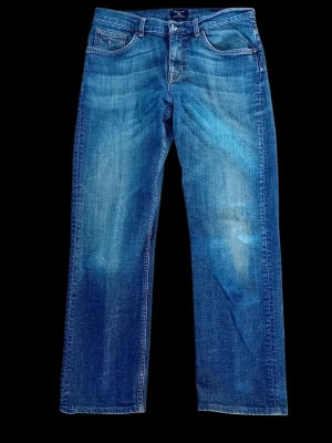 Gant Tyler jeans W33 L32 Herr raka jeans med slitningar - Klassiska blå jeans med raka ben och lätt slitna detaljer framtill. Jeansen har fem fickor, normal midja och stängs med knapp och dragkedja. Materialet är denim i bomull och färgen är mellanblå med ljusare partier. Lite slitage vid en söm (vänster)när de sitter på. Finns i rök- och djurfritt hem.