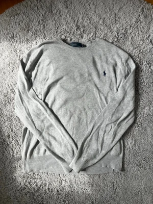 Ljusgrå tröja från Polo Ralph Lauren - Ljusgrå, stickad långärmadtröja från Polo Ralph Lauren med rund halsringning och broderad marinblå logga på bröstet. Tröjan har ribbade muddar vid ärmslut och nederkant, perfekt för en stilren och enkel look. Tröjan är i storlek M men sitter lite tajt. Så jag skulle säga att den mer passar som storlek S. Den har inga hål eller så. Väldigt bra skick!