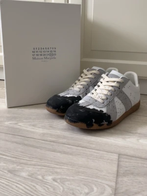 Maison Margiela sneakers med färgstänk - Unika sneakers från Maison Margiela i vitt och grått med svart färgstänk på tå och sidor. Klassisk låg modell med beige snörning och gummisula. Tillverkade i läder och mocka för en lyxig känsla och cool look.