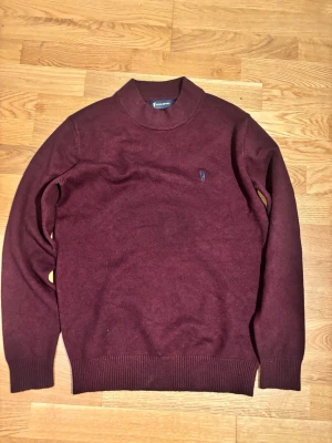 Polo Sport Vinröd Stickad Tröja med Polokrage - En stilren och klassisk stickad tröja från Polo Sport i en djup, vinröd färg. Modellen har en elegant polokrage (mock neck) som ger en sofistikerad look. Perfekt för höst och vinter. 