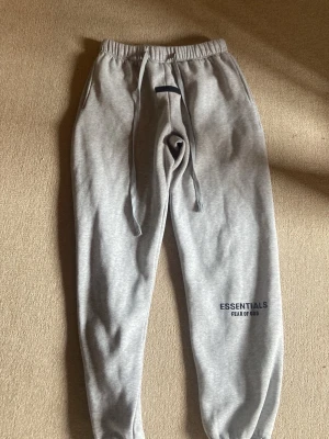 Grå mjukisbyxor Essentials Fear of God - Snygga grå mjukisbyxor från Essentials Fear of God med elastisk midja och dragsko. Byxorna har tryckt logga på benet och muddar vid bensluten. Perfekta för en chill och avslappnad stil.