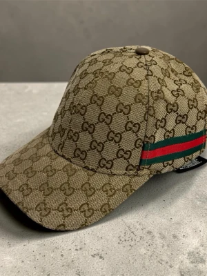 Beige keps från Gucci  - Snygg beige keps från Gucci med klassiskt GG-monogram över hela kepsen och en grön-röd rand på sidan. Justerbar rem bak i brunt läder och böjd skärm. Tillverkad i canvasmaterial som ger en exklusiv känsla.