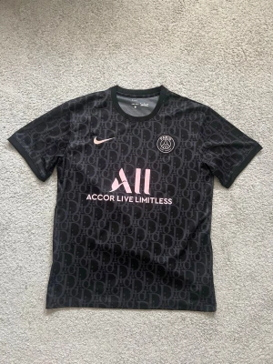 Svart PSG x Nike matchtröja - Snygg svart PSG matchtröja från Nike med diskret cd-mönster över hela tyget. Tröjan har rosa detaljer, klubbmärke på bröstet och texten 'Accor Live Limitless' framtill. Tillverkad i lätt och ventilerande material, perfekt för fotboll eller träning.