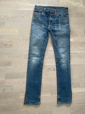 Nudie jeans - Snygga blå jeans med smal passform och slitna detaljer på framsidan och baksidan. Klassisk femficksmodell med dragkedja och knapp. Jeansen har slitsar vid bensluten och kontrastsömmar. Perfekta för en avslappnad och trendig look. Low waist, unisex, några skavanker går att se om man kollar noga.