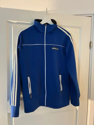 Blå Adidas track jacket med vita ränder - Säljer en blå adidas track jacket i storlek M, den är väldigt liten i storlek samt lite nopprig.