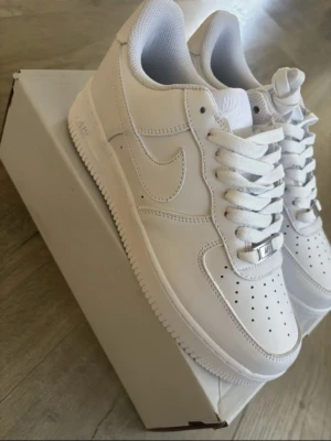 Nike Air Force 1  - Klassiska Nike Air Force 1 sneakers i helvitt läder med perforerad tåbox, vit sula och vita skosnören. Ikonisk Swoosh-logga på sidan och diskret Air-detalj på sulan. Perfekt för dig som gillar stilrena och tidlösa sneakers. Helt nya!!!