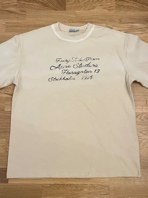 Beige t-shirt från Acne Studios - Beige t-shirt från Acne Studios med svart text på bröstet i handskriven stil. Klassisk rund halsringning och avslappnad passform. Perfekt för en stilren och trendig look.