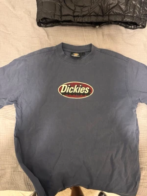 Blå Dickies t-shirt med logga - Säljer en blå t-shirt från Dickies med klassisk logga tryckt på bröstet. T-shirten har rund hals och korta ärmar, perfekt för en avslappnad och streetinspirerad stil. Tillverkad i mjuk bomull för skön komfort.