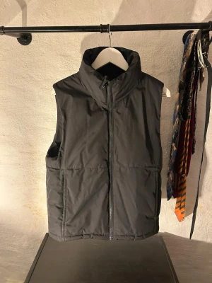Svart dunväst med hög krage - Snygg svart dunväst med hög krage och dragkedja framtill. Västen har en stilren design utan synliga loggor och två sidofickor. Perfekt att slänga över en hoodie eller tröja för extra värme under kyliga dagar.