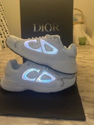 Vita Dior sneakers med reflekterande CD - Snygga vita sneakers från Dior med reflekterande CD-logga på sidorna och bak. Skorna har en chunky sula och är tillverkade i mesh och läder för en modern look. Perfekta för dig som vill sticka ut med exklusiva detaljer och trendig design.