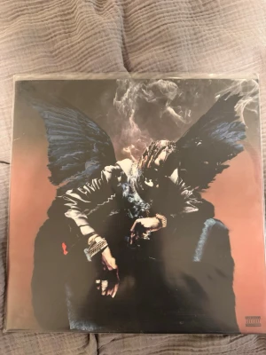 Travis scott beirbs in the trap - Säljer ett albumomslag med ett konstnärligt motiv på framsidan där en person med svarta vingar sitter i rök. Baksidan visar en örn ovanför en brinnande banderoll med låttitlar. Färgerna är mörka med inslag av blått, svart och orange.