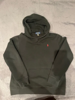 Svart hoodie från Polo Ralph Lauren - Säljer en svart hoodie från Polo Ralph Lauren med klassisk röd logga broderad på bröstet. Hoodien har huva, känguruficka och ribbade muddar vid ärmar och nederkant. Tillverkad i mjukt bomullsmaterial som är skönt att bära.