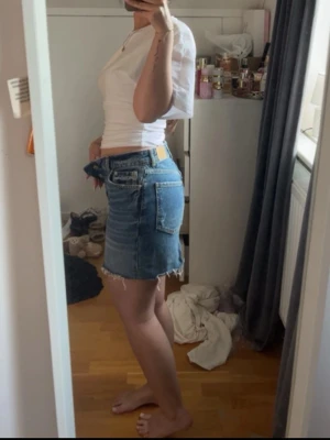Blå jeanskjol med fransig kant - Säljer en blå jeanskjol i kort modell med rå, fransig kant nertill. Kjolen har klassiska fem fickor, knappgylf och en lätt tvättad look. Perfekt för dig som gillar en avslappnad och trendig stil.