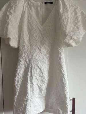 Vit kort klänning med puffärm - Superfin vit kort klänning med struktur och puffärmar. Klänningen har v-ringning och är figurnära med en snygg textur i tyget. Perfekt för sommarens tillfällen och studentfirande.