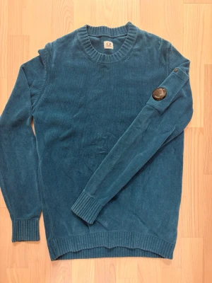 Blå stickad tröja från C.P. Company - Säljer en blå stickad tröja från C.P. Company med rund halsringning och ribbade muddar. Tröjan har en ikonisk svart linsdetalj på ena ärmen som ger en unik look. Perfekt för dig som gillar streetwear och snygga detaljer.