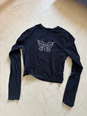 Svart långärmad topp med fjäril - Snygg svart långärmad topp med ett stort fjärilsmotiv i silvriga strasstenar på bröstet. Croppad modell med rund halsringning. Perfekt för dig som gillar trendiga detaljer och vill sticka ut lite extra.
