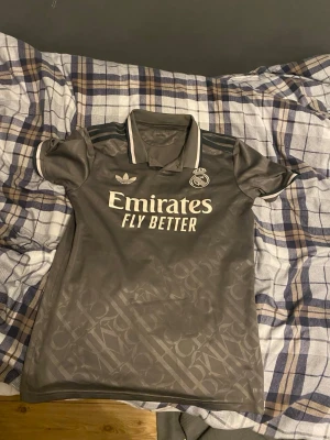 Grå Real Madrid matchtröja Adidas - Snygg grå Real Madrid matchtröja från Adidas med vita detaljer och klubbmärke på bröstet. Tröjan har korta ärmar, krage med vit kant och diskret mönster i tyget. Perfekt för fotboll eller träning.