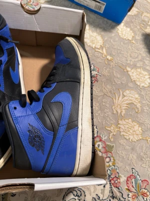 Air Jordan 1 Nike blå och svart  - Snygga höga sneakers i svart och blått med klassisk siluett och tydlig swoosh på sidan. Skorna har svarta skosnören, blå detaljer och svart yttersula. Perfekta för dig som gillar streetstyle och vill sticka ut med en ikonisk look.