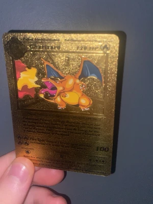 Guldfärgat Charizard Pokémonkort - Säljer ett unikt guldfärgat Pokémonkort med Charizard-motiv. Kortet är tillverkat i metall och har en glittrig finish med färgglad bild på Charizard som sprutar eld. Baksidan har Pokémon-loggan i relief. Perfekt för samlare eller Pokémonfans.