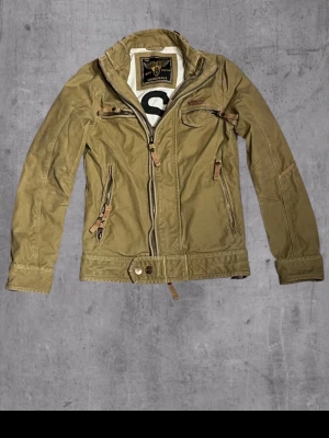 Beige vindjacka med dragkedjor - Snygg beige vindjacka med flera dragkedjor och detaljer i läderimitation. Jackan har hög krage, lång ärm och praktiska fickor framtill. Perfekt för dig som gillar en sportig och avslappnad stil. Passformen är normal och materialet känns slitstarkt.