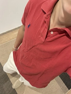 Röd pikétröja från Polo Ralph Lauren - Klassisk röd pikétröja från Polo Ralph Lauren med blå logga broderad på bröstet. Tröjan har krage, knappar vid halsen och är tillverkad i bomull. Custom fit-modell som ger en snygg passform och passar perfekt till både jeans och chinos. Finns små defekter vid öppningarna vid midjan.