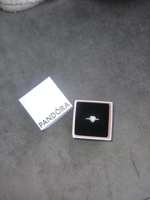 Silverring med hjärta från Pandora - Säljer en snygg silverring från Pandora med ett glittrande hjärta i mitten och små stenar längs bandet. Ringen kommer i original ask och har en elegant och romantisk design som verkligen sticker ut.