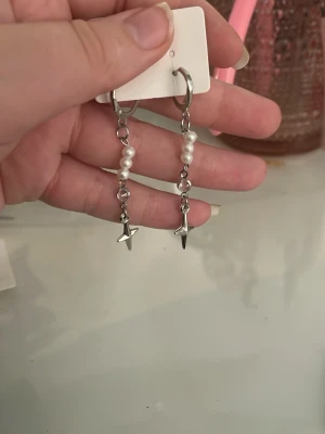 Silvriga örhängen med pärlor och stjärnor - Säljer ett par snygga silvriga örhängen med hängande kedjor, vita pärlor och små stjärnberlocker längst ner. Örhängena har en modern och trendig design som passar perfekt till dig som gillar unika accessoarer.