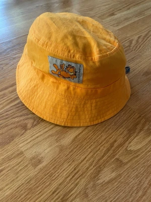 Gul bucket hat med Garfield-tryck - Cool bucket hat i starkt gul färg med ett broderat Garfield-märke framtill. Hatten är i mjukt tyg och har klassisk form med nedvikt brätte. Perfekt för att sticka ut i mängden och ge din outfit en lekfull touch.