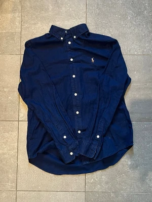 Mörkblå Ralph lauren skjorta  - Säljer en klassisk mörkblå skjorta med vita knappar och button down-krage. Skjortan har lång ärm och en liten broderad logga på bröstet. Tillverkad i bomull för en skön känsla och snygg passform. Det står 14-16 jag skulle uppskatta att det är xs-s