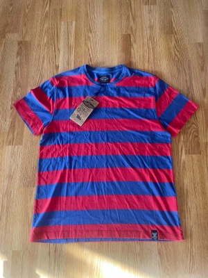 Randig t-shirt från DePalma - Säljer en randig t-shirt från DePalma i blått och rött. T-shirten har klassisk passform, rund hals och korta ärmar. Tillverkad i mjuk bomull och har en snygg streetwear-vibe. Perfekt för dig som gillar färg och mönster.