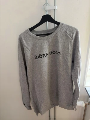 Grå sweatshirt från Björn Borg - Snygg grå sweatshirt från Björn Borg i storlek XXL. Tröjan har rund halsringning, långa ärmar och svart tryck med loggan på bröstet. Tillverkad i mjukt material som känns skönt mot huden. Perfekt för en avslappnad och sportig stil.