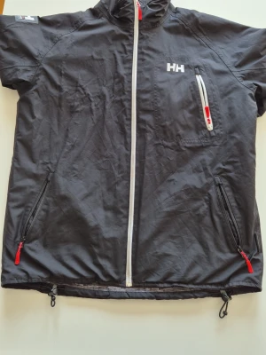 Svart jacka från Helly Hansen - Snygg svart höstjacka från Helly Hansen med vit dragkedja och röda detaljer. Jackan har tre fickor med dragkedja, hög krage och justerbar nederkant. Perfekt för blåsiga dagar och enkel att packa ner i väskan.