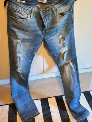 Blå slitna skinny jeans med hål - Säljer ett par blå skinny jeans med slitningar och stora hål på båda benen. Jeansen har klassisk femficksmodell, låg midja och är tillverkade i denim. Perfekta för en avslappnad och trendig look.