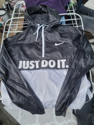 Nike svartvit vindjacka med huva - Nike vindjacka i svart och vitt med huva och halv dragkedja. Stor 'JUST DO IT.'-text framtill och klassisk Nike-logga på bröstet. Jackan har dragsko i huvan och elastiska muddar. Perfekt för blåsiga dagar och streetwear-stil.