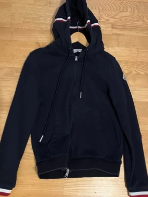 Svart Moncler hoodie med dragkedja - Snygg svart hoodie från Moncler med hel dragkedja, huva med vita och röda ränder samt matchande detaljer på ärmsluten. Klassisk Moncler-logga på ena ärmen och praktiska fickor framtill. Perfekt för en stilren och sportig look.