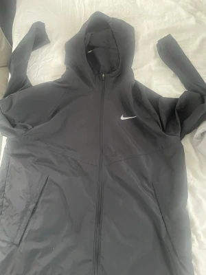 Svart Nike vindjacka med huva - Snygg svart vindjacka från Nike med huva och dragkedja framtill. Jackan har två sidofickor med dragkedja och en vit Nike-logga på bröstet. Perfekt för löpning eller när du vill ha en sportig look. Tillverkad i lätt och vindtätt material.