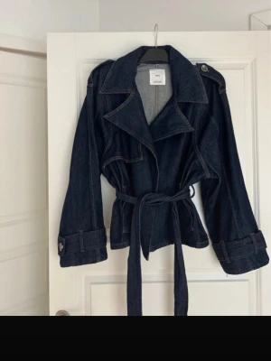 Mörkblå denim jeansjacka med skärp - Snygg mörkblå jeansjacka med knytskärp i midjan och detaljer. Jackan har markerad midja för en cool siluett. Perfekt för dig som gillar en trendig och avslappnad look.