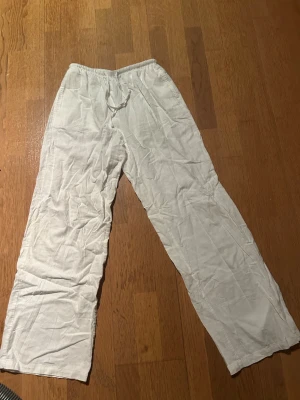 Vita linnebyxor med knyte  - Säljer ett par vita linnebyxor med avslappnad passform och knyte i midjan. Byxorna har raka ben och är tillverkade i ett lätt och luftigt linnematerial, perfekta för en chill och stilren look.