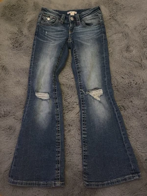Blå bootcut jeans med slitningar - Snygga blå bootcut jeans med slitna detaljer på knäna och ljusare tvätt framtill. Jeansen har fem fickor, knappgylf och dekorativa sömmar på bakfickorna. Perfekta för en avslappnad och trendig look.